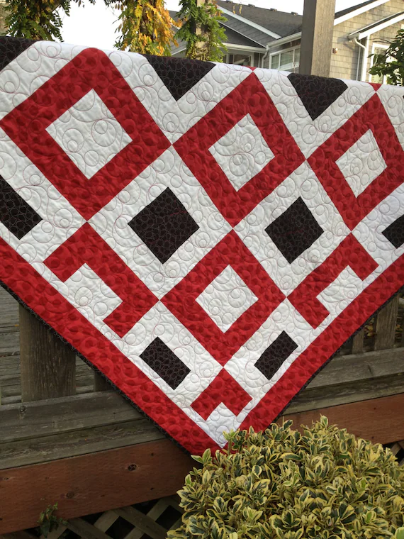 Red, white, and dark patchwork quilt with geometric square pattern draped over a wooden railing outdoors, bold warm color blocking