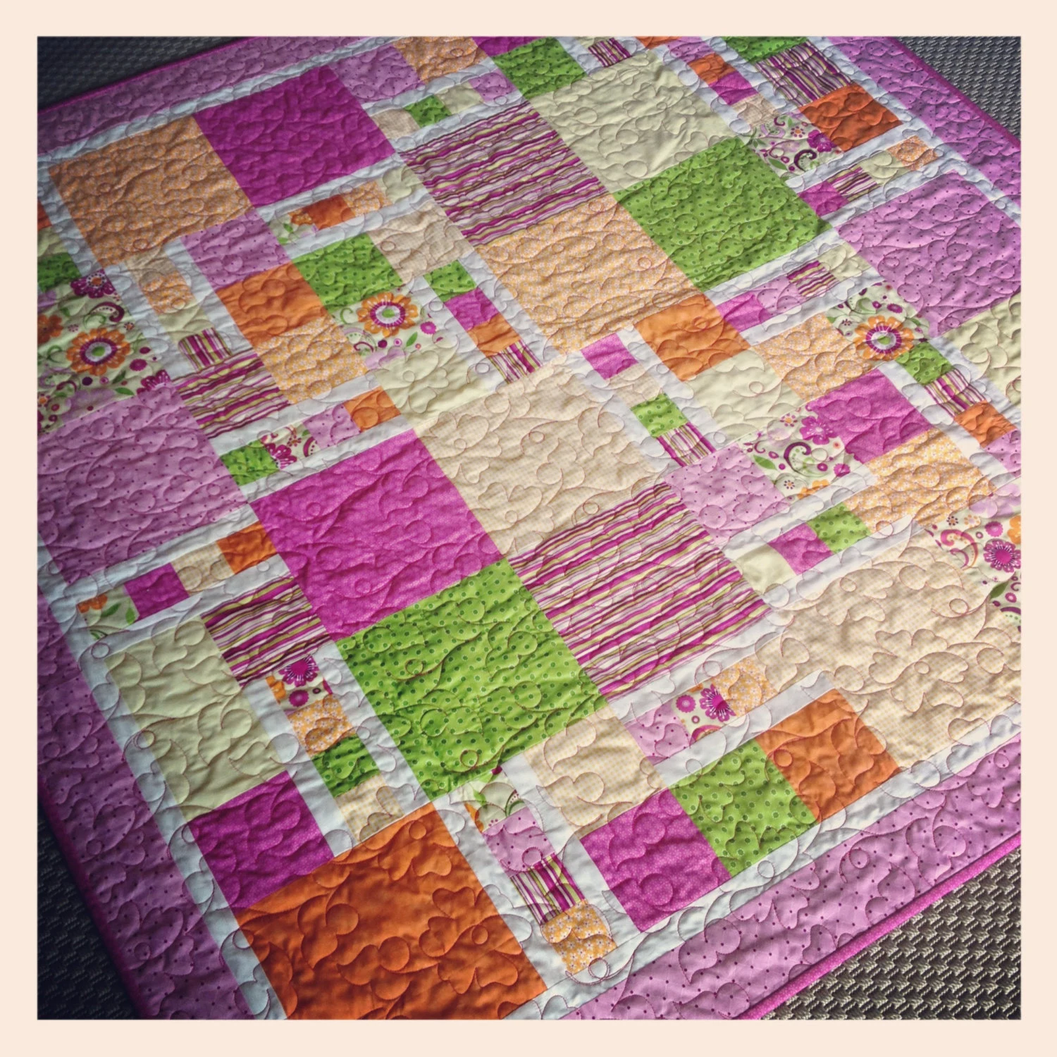 A patchwork quilt with pink, orange, green, and cream squares arranged in a block pattern, decorative quilting stitches visible across the surface.