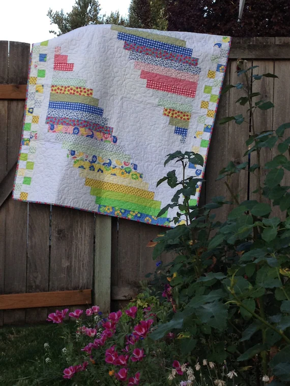 Patchwork quilt with colorful striped bargello-style design and checkered border hanging on a wooden fence near flowers, warm and inviting