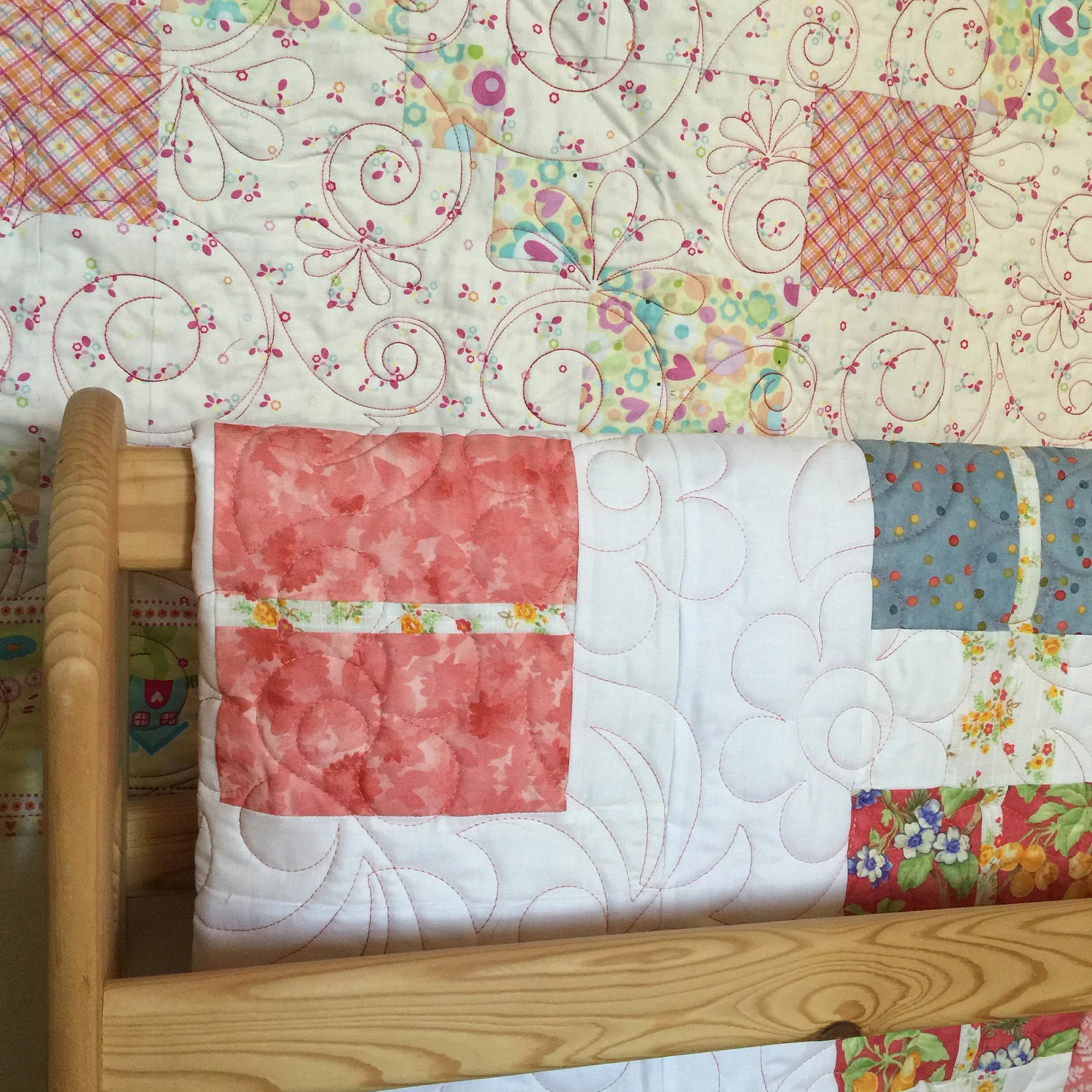 Close-up of a patchwork quilt with pastel floral and plaid squares and decorative swirl quilting stitches, draped over a soft surface in muted tones