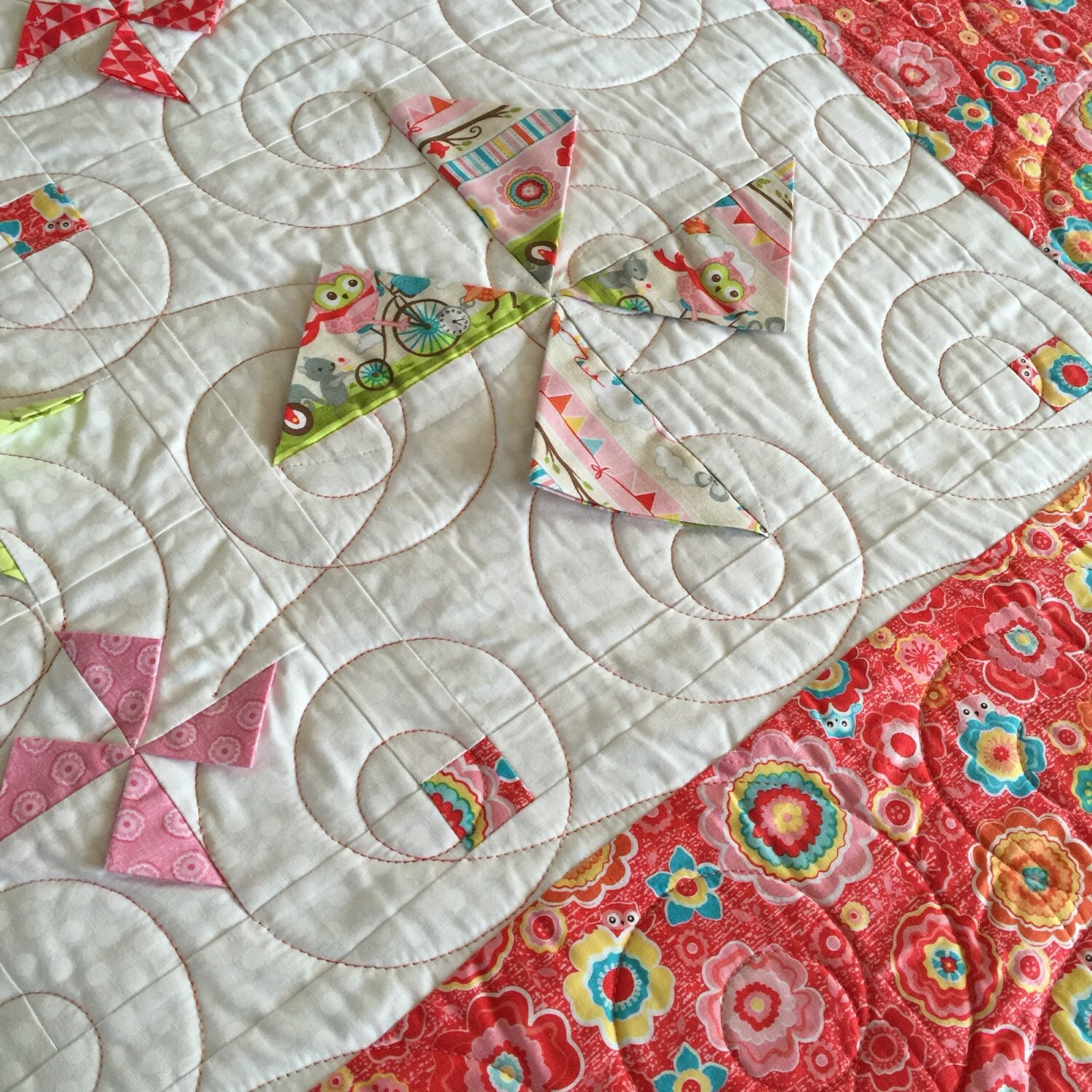 Close-up of white quilted fabric with swirl stitching and small pinwheel-shaped fabric accents in pink, green, and red, soft and friendly mood.