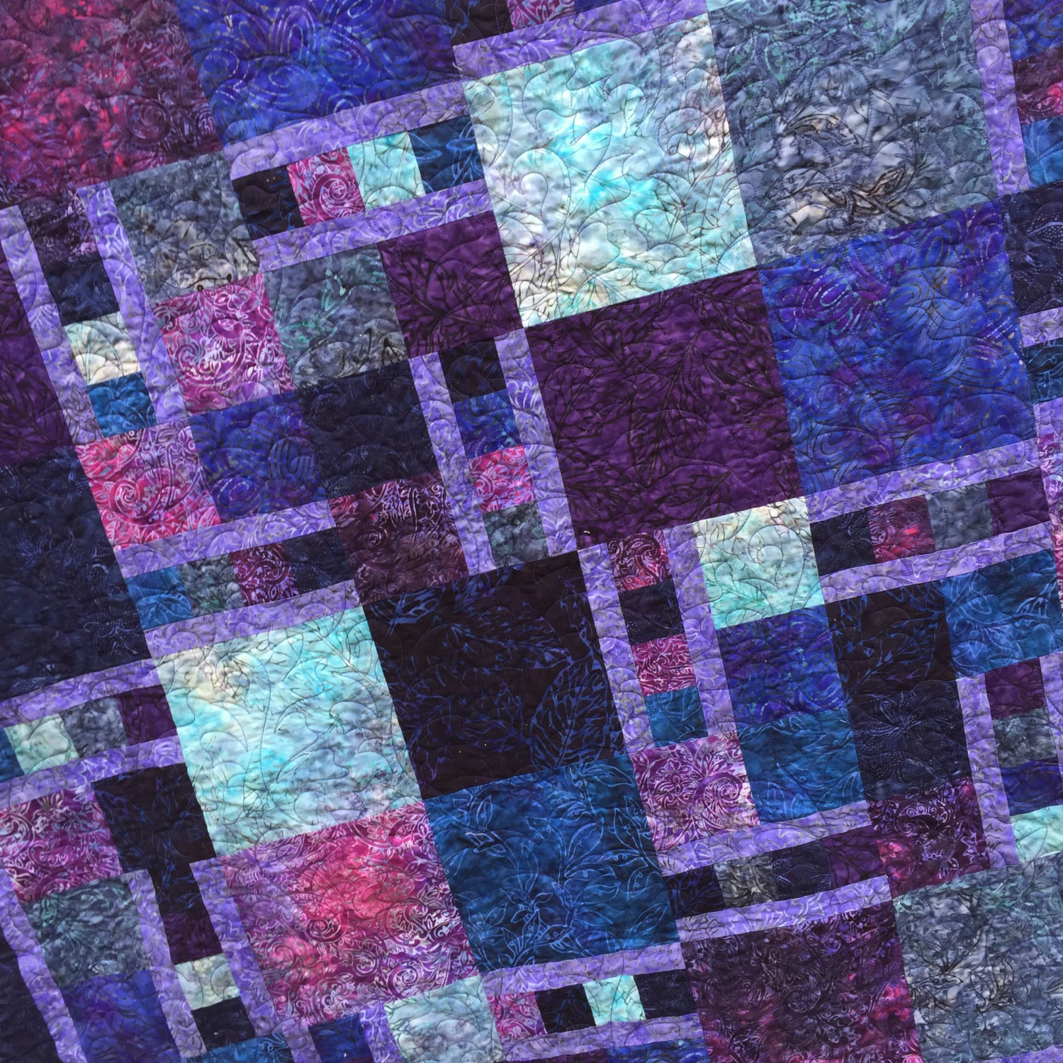 Patchwork quilt in deep purple, blue, teal, and magenta batik fabrics arranged in a block pattern, soft stitched lines, calm creative mood.
