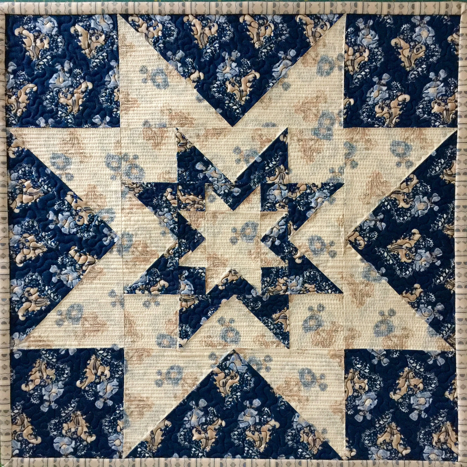 A square quilted block featuring a navy blue and cream star pattern with floral print fabric, bordered by striped tan fabric, classic mood.