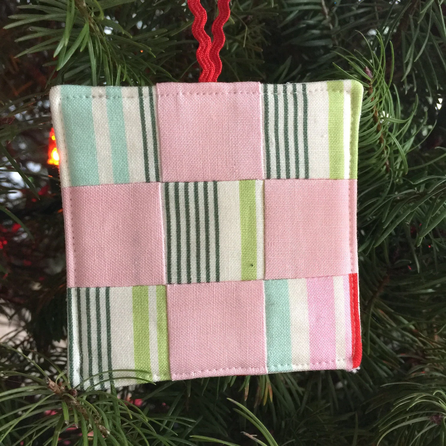 A small patchwork fabric ornament in pink, green, and white striped squares, hanging by a red cord, cheerful holiday mood.